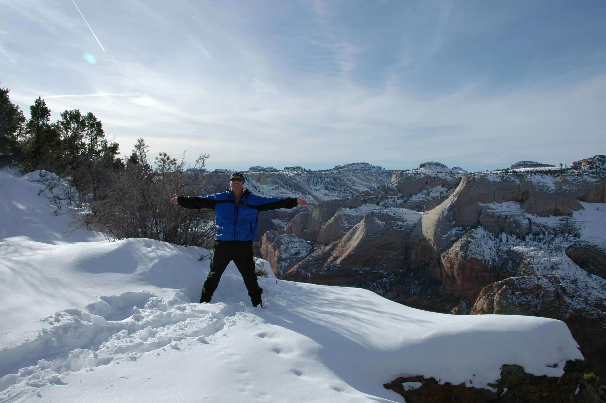 A person stands joyfully on a snowy ledge surrounded by mountains.