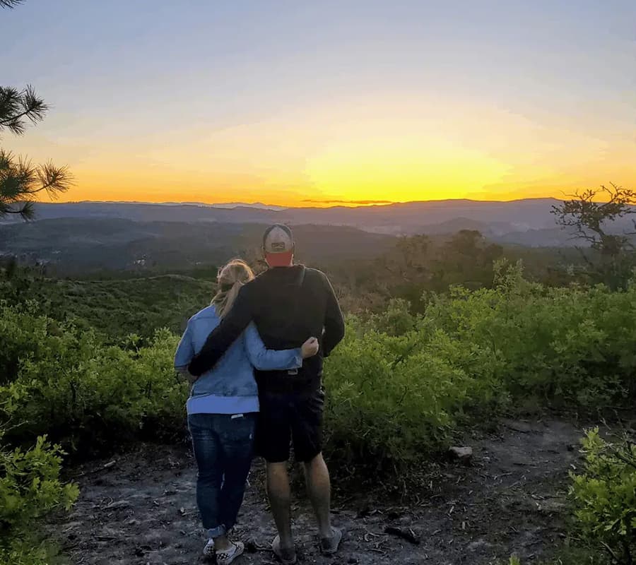 A couple stands together, embracing, as they watch a sunset over a lush landscape.