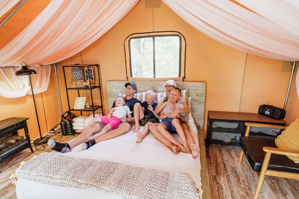 A group of four children and one adult relax on a large bed inside a tent with soft decor.
