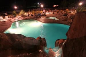 A glowing swimming pool surrounded by lounge chairs and landscaped greenery at night.