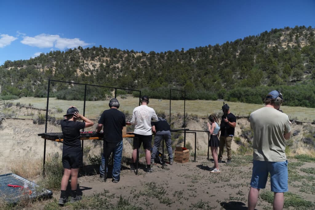 A group of people practice shooting at targets in an outdoor shooting range surrounded by hills.