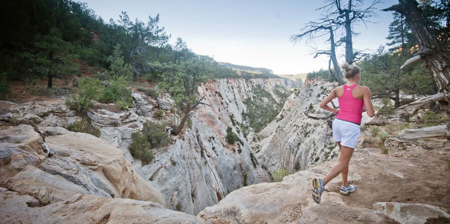 A runner in a pink tank top navigates rocky terrain near a deep canyon surrounded by trees.