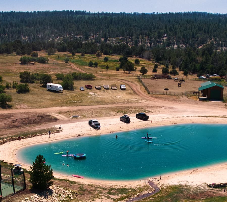 Aerial view of a serene pond surrounded by greenery, with people paddleboarding and parked vehicles nearby.