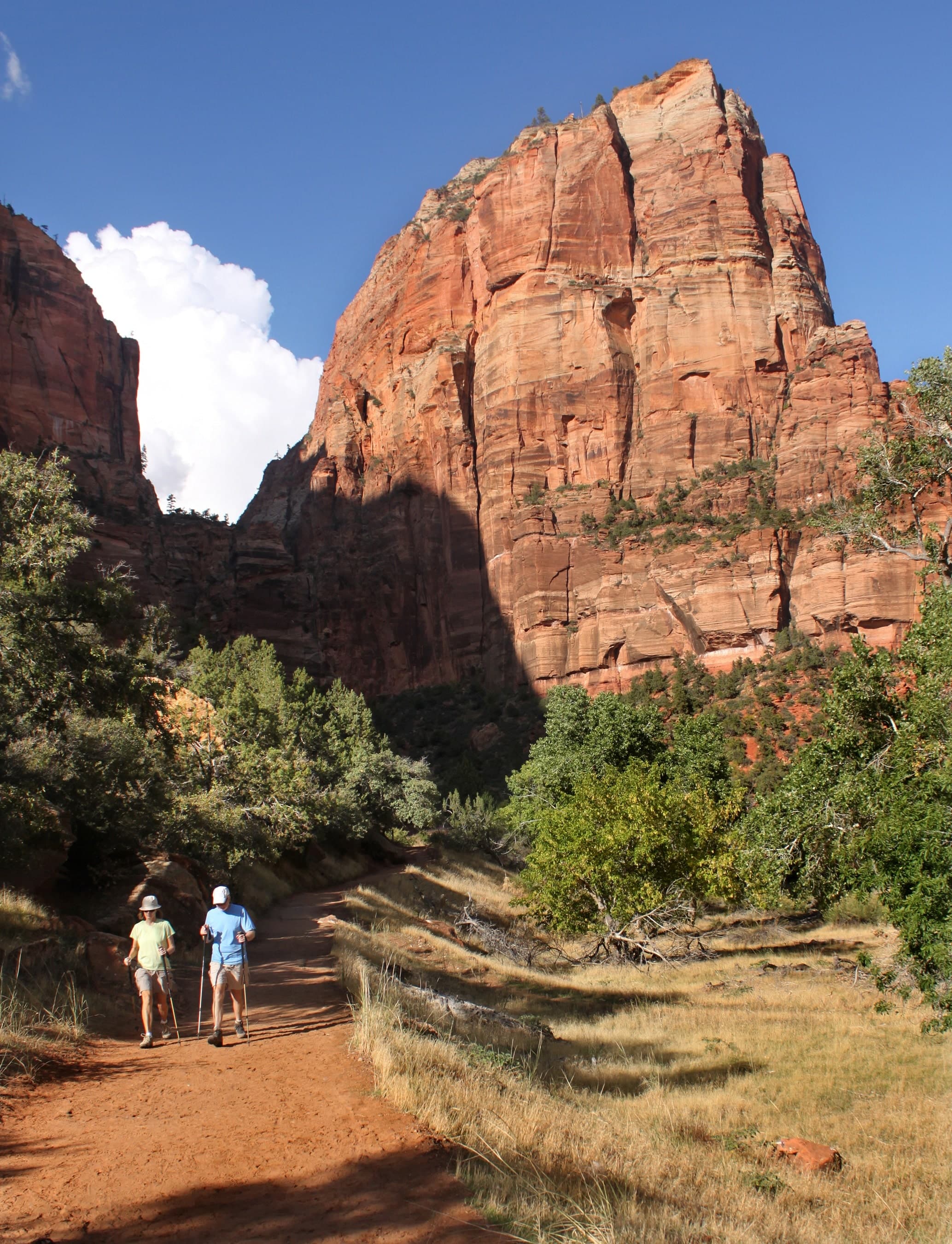 Two hikers walking on a dirt trail surrounded by towering red rock formations and greenery.