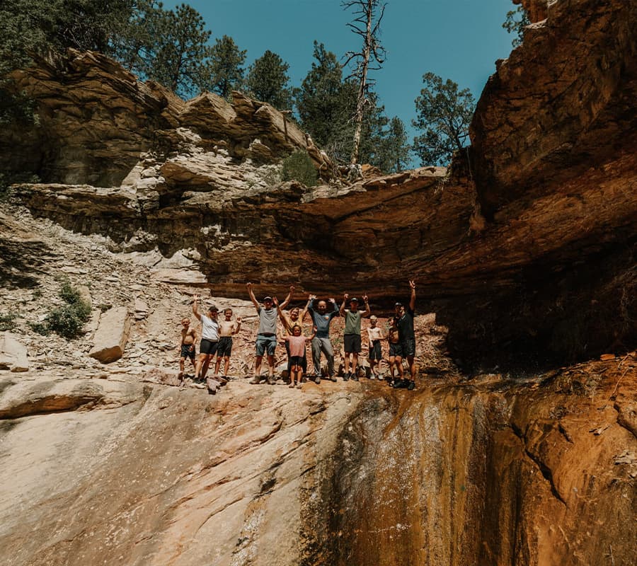 A group of ten people joyfully pose with their arms raised on a rocky ledge in a natural setting.