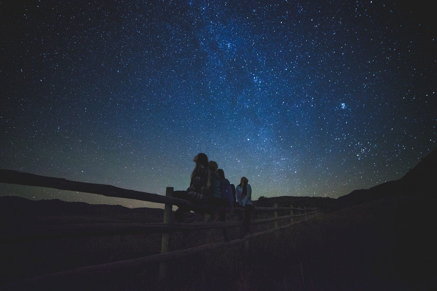 Silhouettes of four people sitting on a fence under a starry night sky.