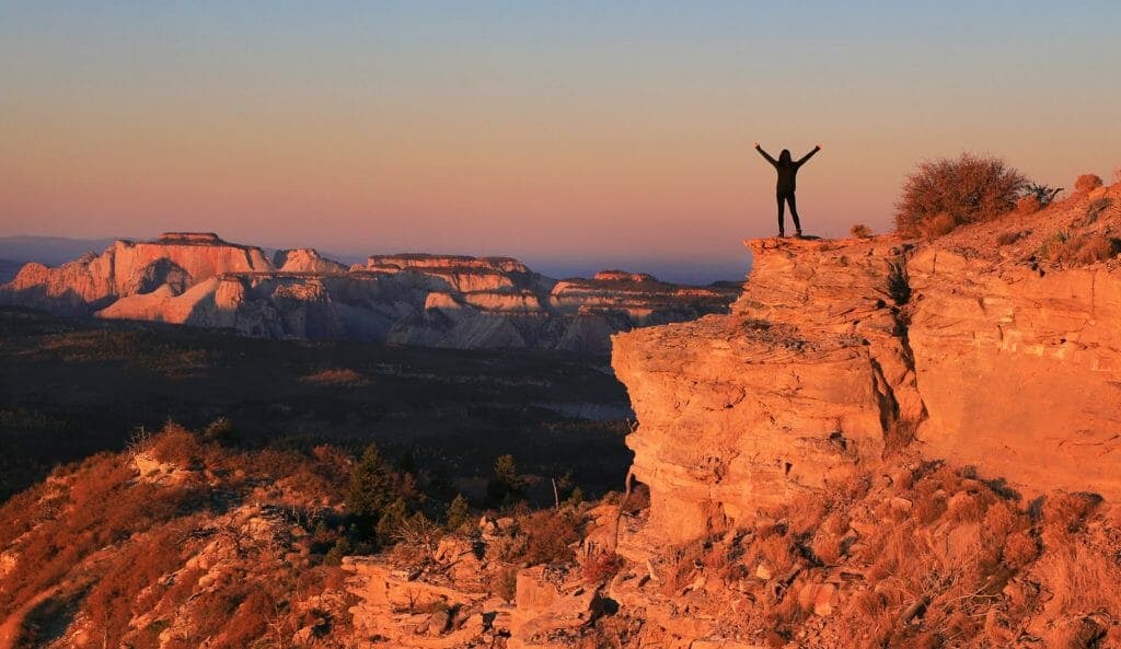 A silhouette of a person with arms raised stands on the edge of a rocky cliff at sunset, overlooking a mountainous landscape.