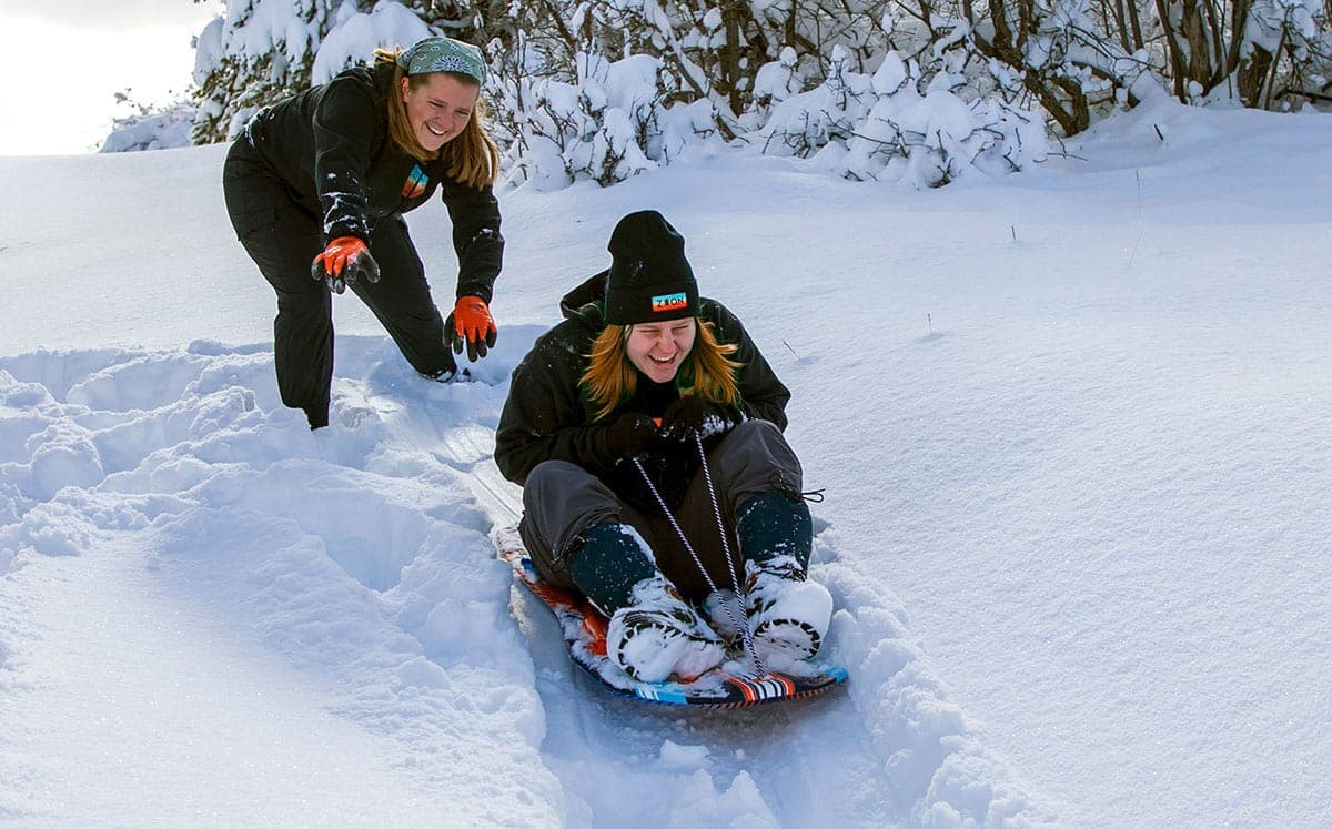 Two people enjoy sledding down a snowy hill, with one person pushing from behind.