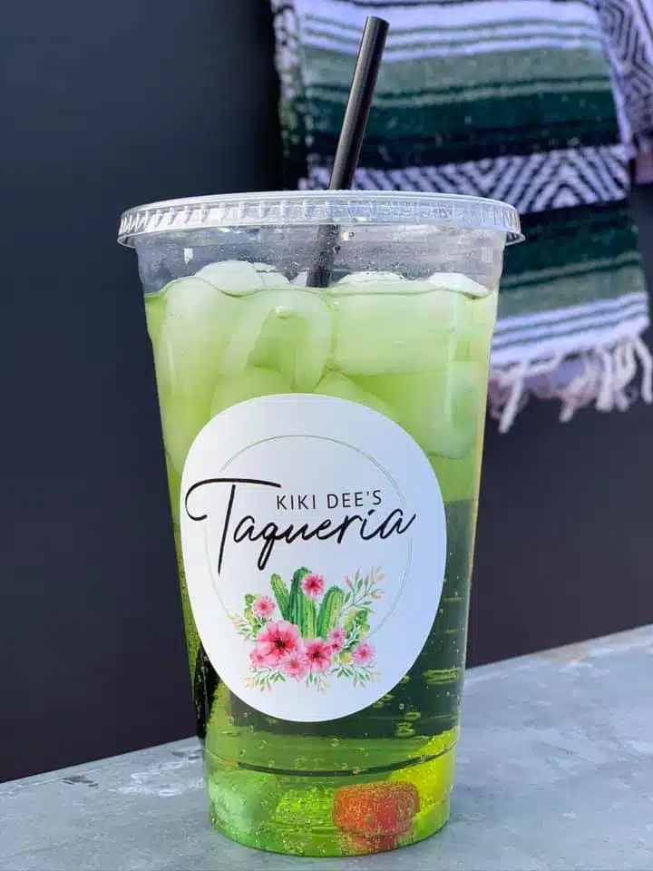 A clear plastic cup filled with a green beverage and ice, featuring a label that reads "KIKI DEE'S Taqueria."