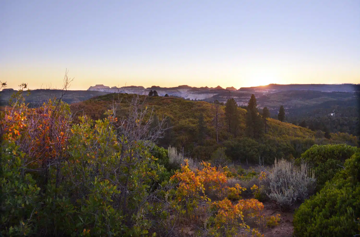 A serene landscape at sunset, featuring rolling hills and a mix of green and orange foliage.