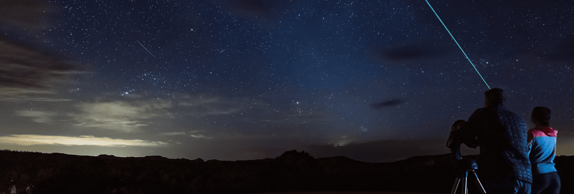 A person points a laser at the starry night sky while another observes through a telescope.