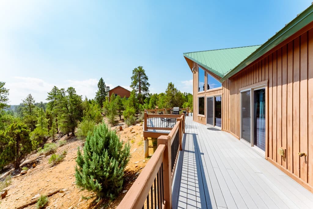 A wooden deck with a green roofed house overlooks a sunlit forested landscape.