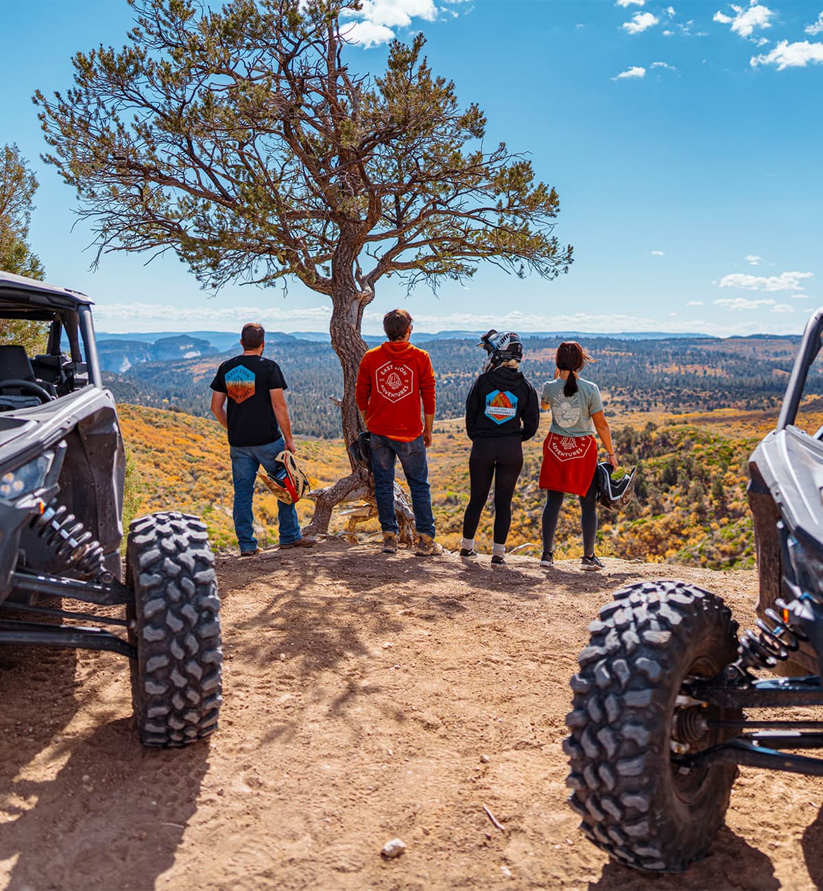 Four individuals gaze at a scenic landscape from a rocky outcrop, with off-road vehicles partially visible in the foreground.