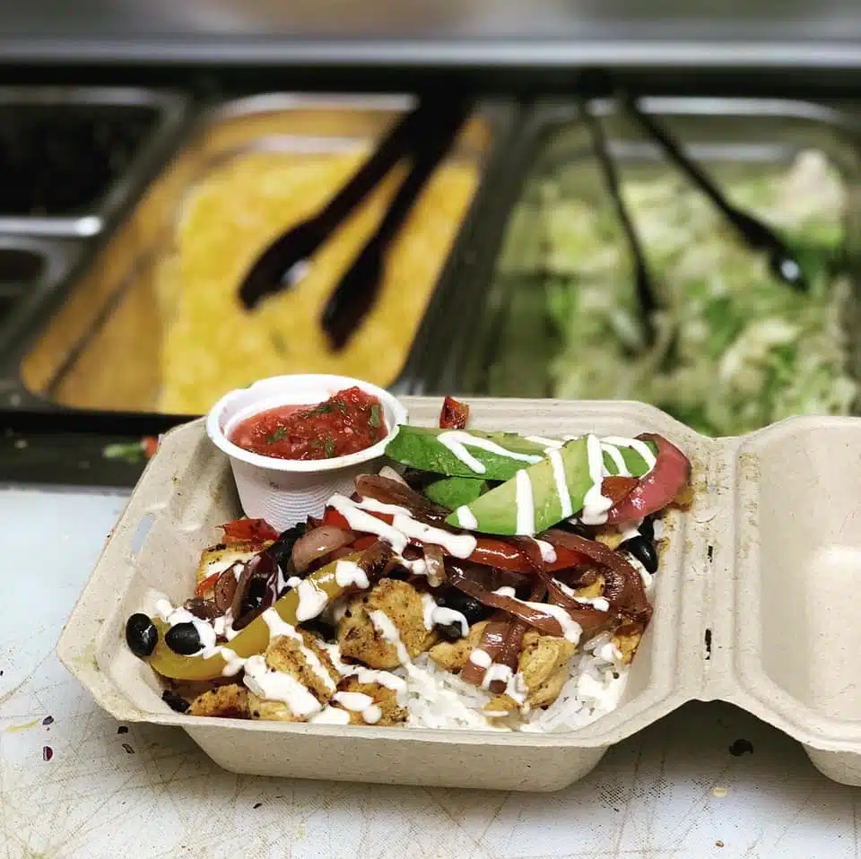 A takeout container filled with rice, grilled chicken, black beans, sautéed peppers, avocado, and a side of salsa.
