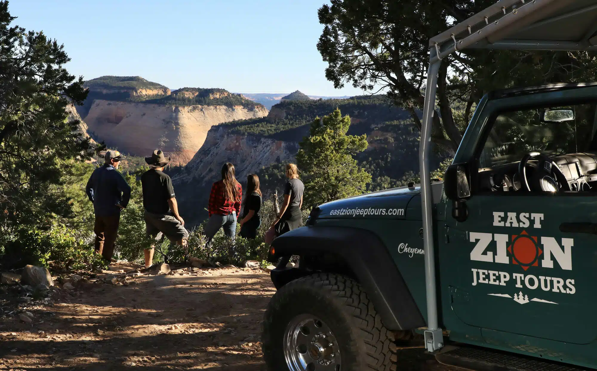 A group of people stands on a trail, overlooking a scenic canyon with a Jeep parked nearby.