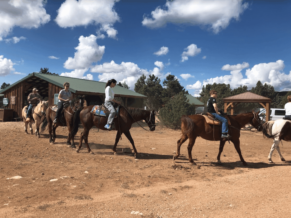 A group of people riding horses along a dirt path near a stable under a partly cloudy sky.