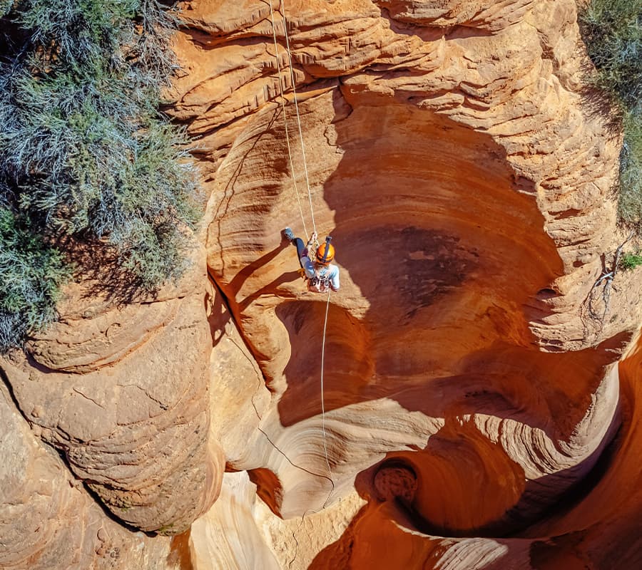 A person rappels into a deep, winding canyon with reddish rock formations.