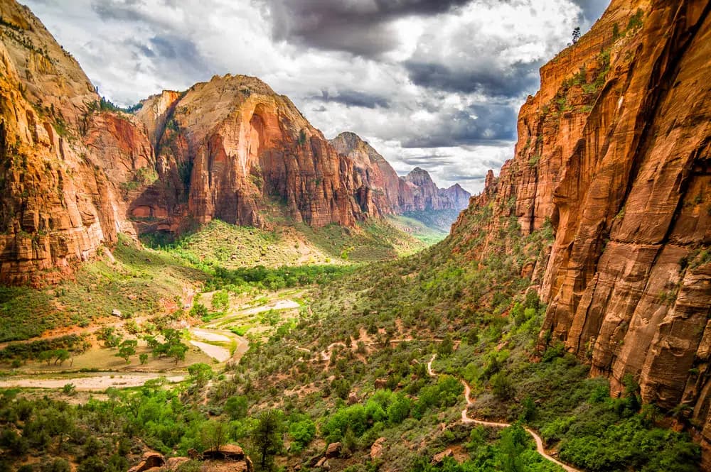 A scenic view of Zion National Park featuring towering red rock cliffs and a lush green valley under cloudy skies.