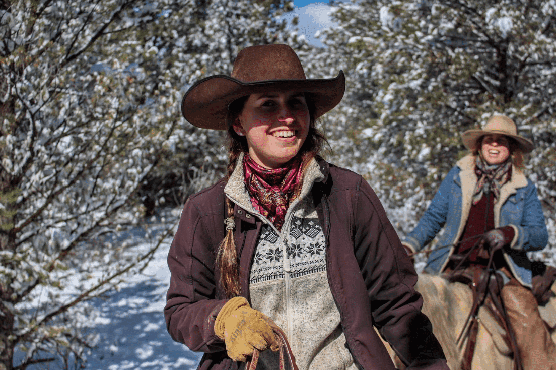 Two cowgirls riding horses through a snowy landscape, smiling in the sunlight.