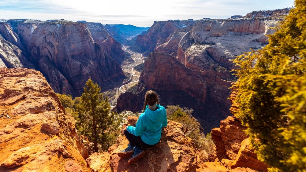 A person sits on a rocky ledge overlooking a vast canyon landscape.