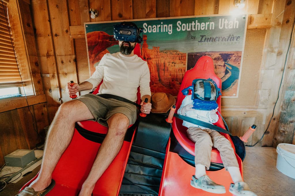 A man and a child sit in red virtual reality seats, both wearing VR headsets and holding controllers.
