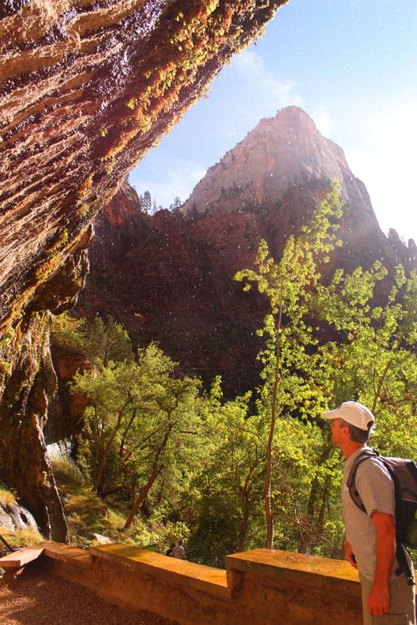 A hiker stands under a rocky overhang, gazing up at a mountain surrounded by greenery and sunlight.
