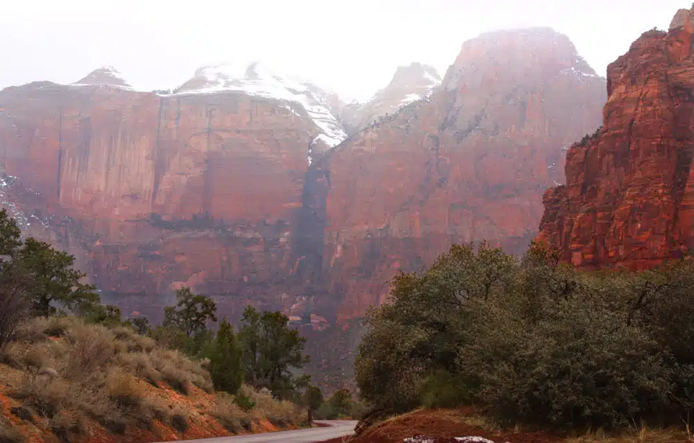 A winding road leads through a canyon with towering, snow-capped red rock cliffs.