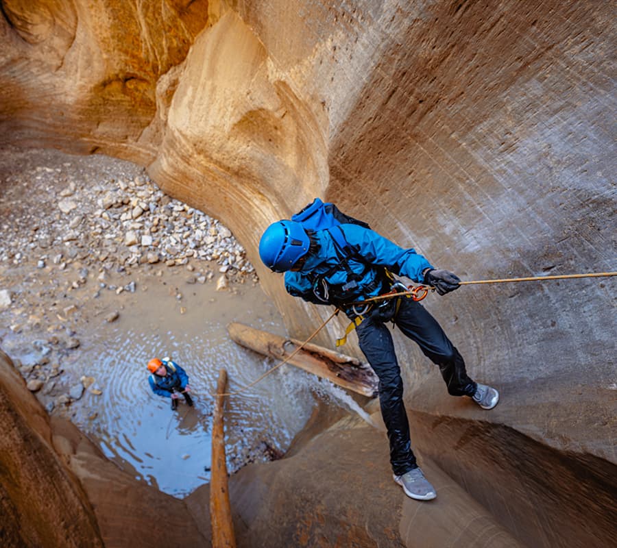 Two climbers navigate a narrow canyon, one rappelling down while the other stands in water below.