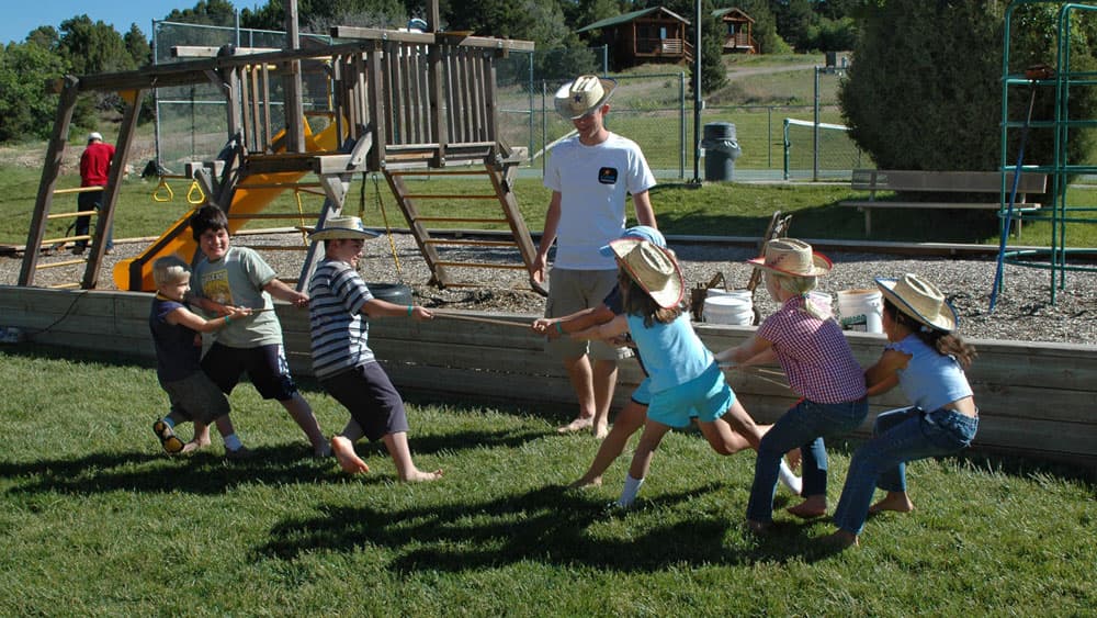 Children in cowboy hats playing tug-of-war on a sunny day at a playground.