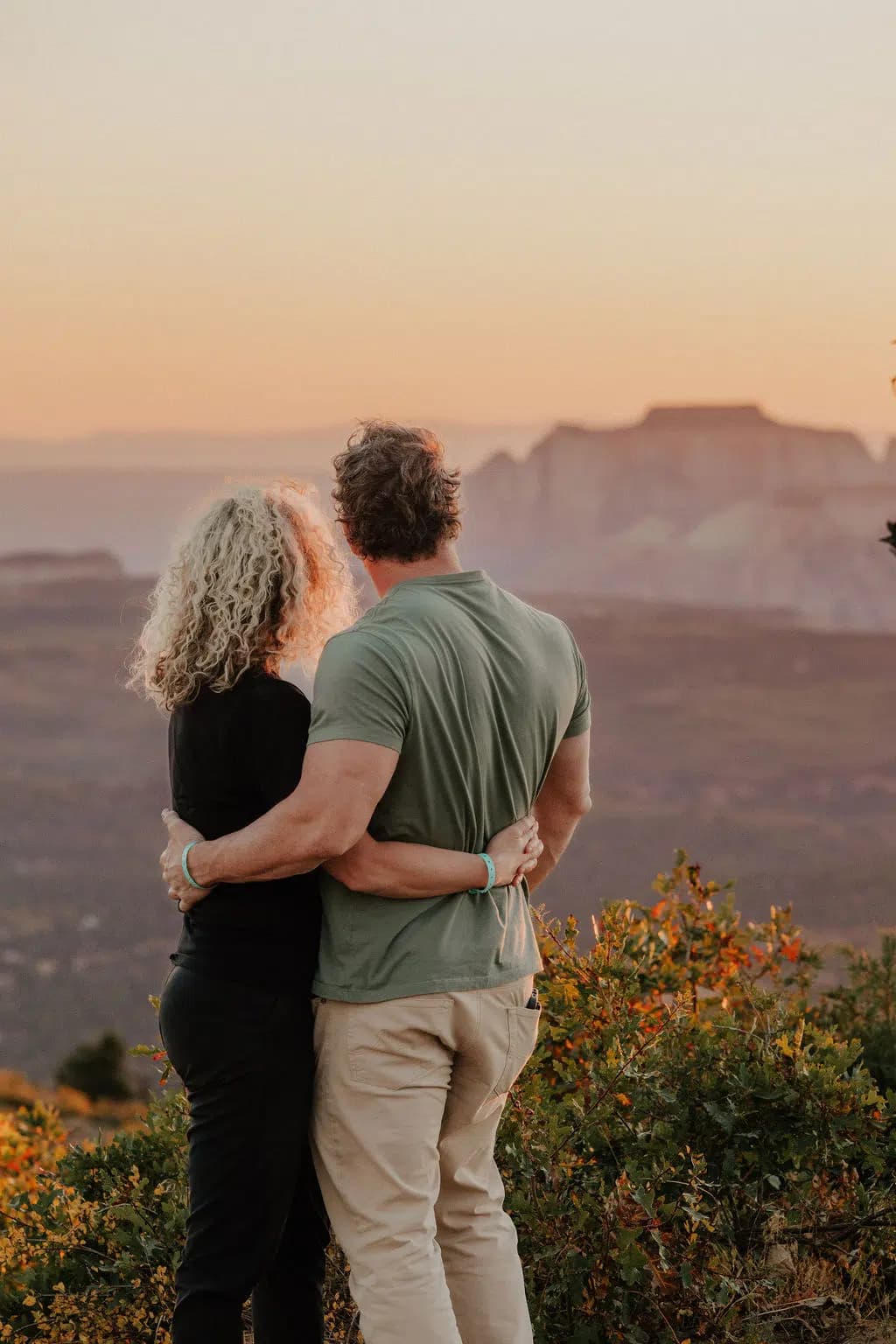 A couple embraces while watching a sunset over a mountainous landscape.