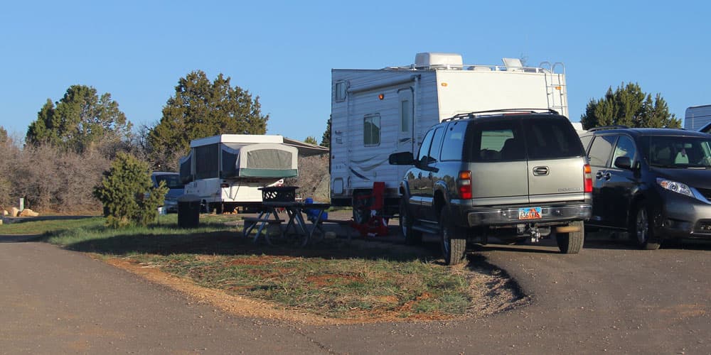 A camper and an RV parked in a campsite with nearby picnic tables.