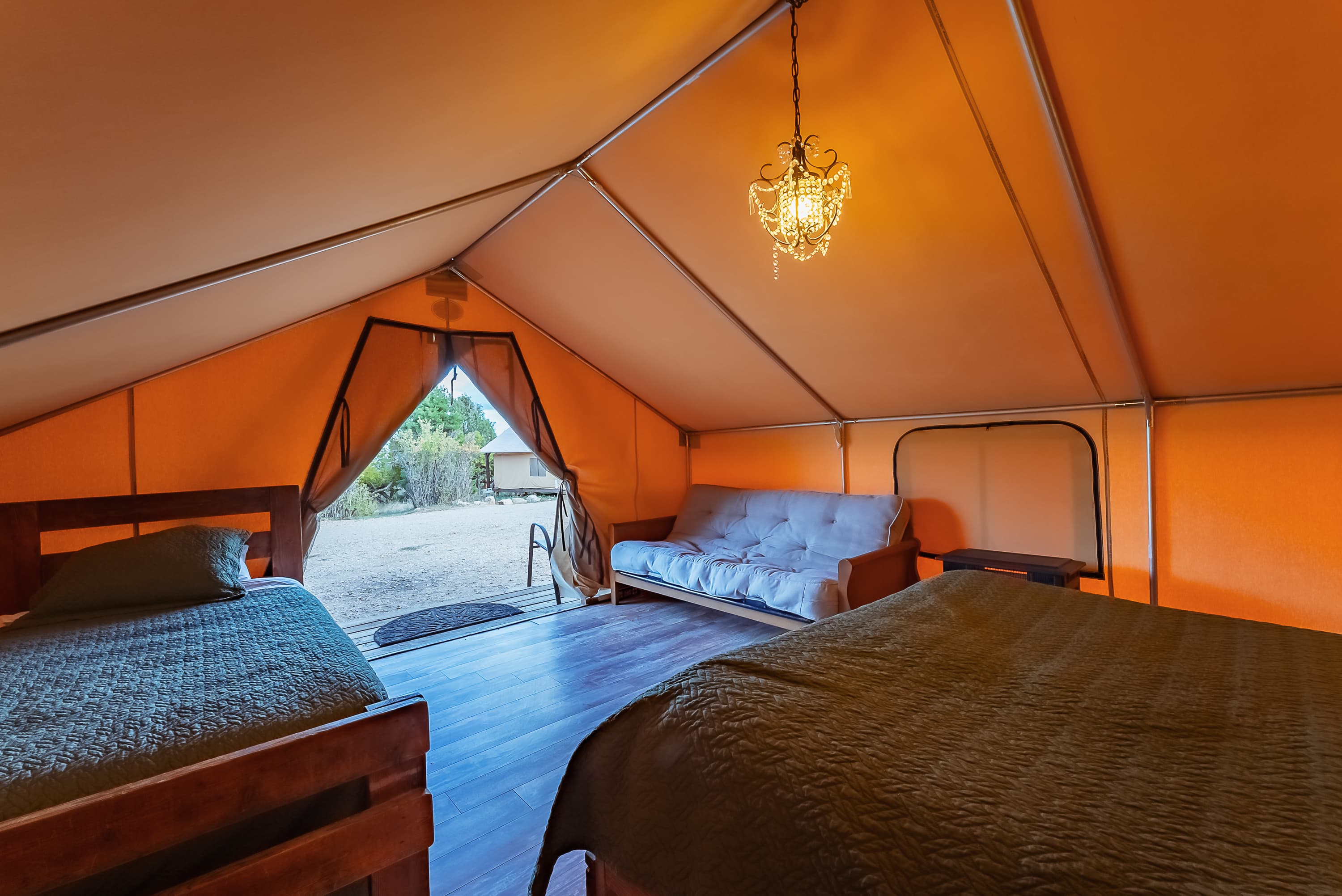 Interior of a cozy orange canvas tent featuring two beds and a sofa, with a chandelier and an open entrance to the outdoors.