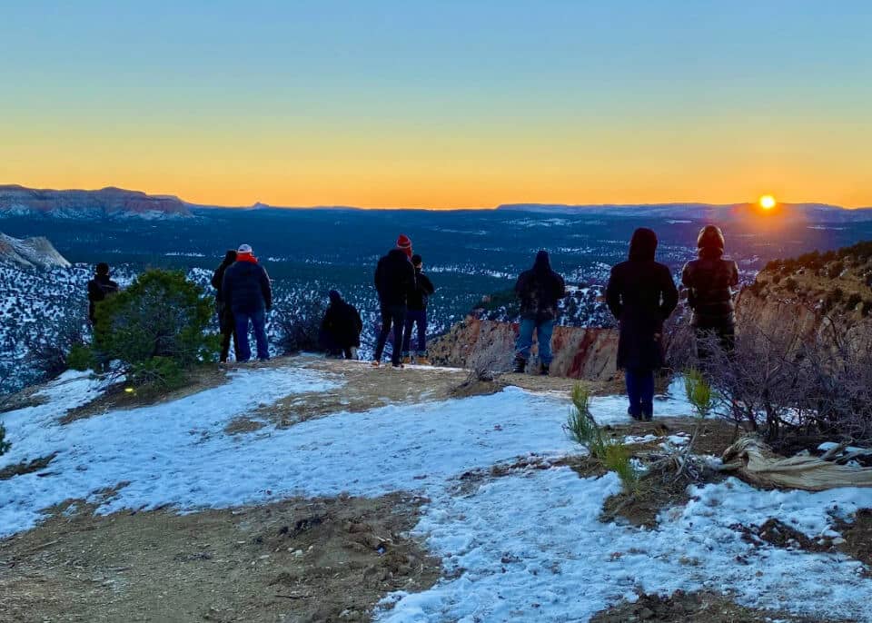 A group of people standing on a snowy cliff at sunrise, admiring the landscape.