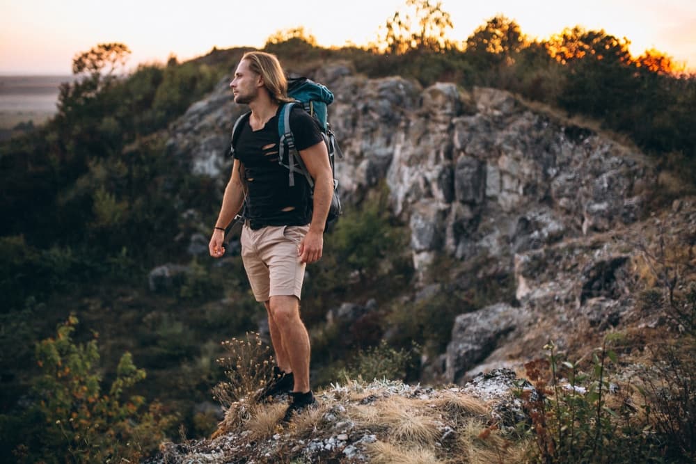 A young man stands on a rocky ledge, gazing into the distance with a backpack on his back, surrounded by nature at sunset.
