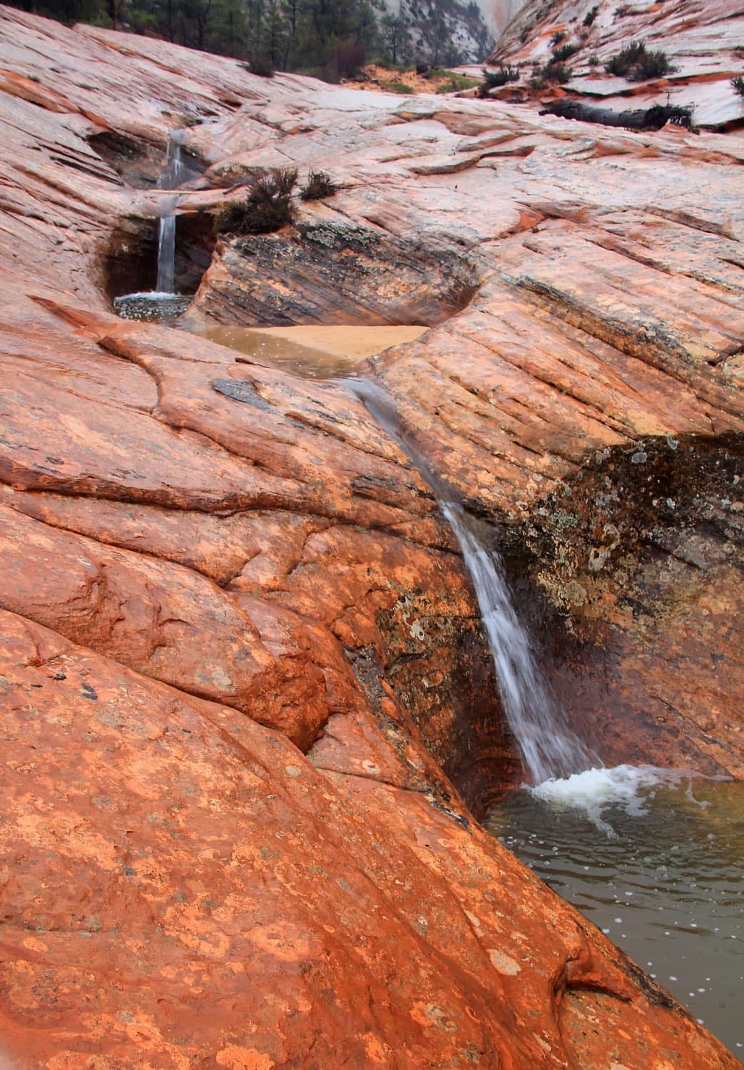 A cascading stream flows over reddish rock formations into a small pool.