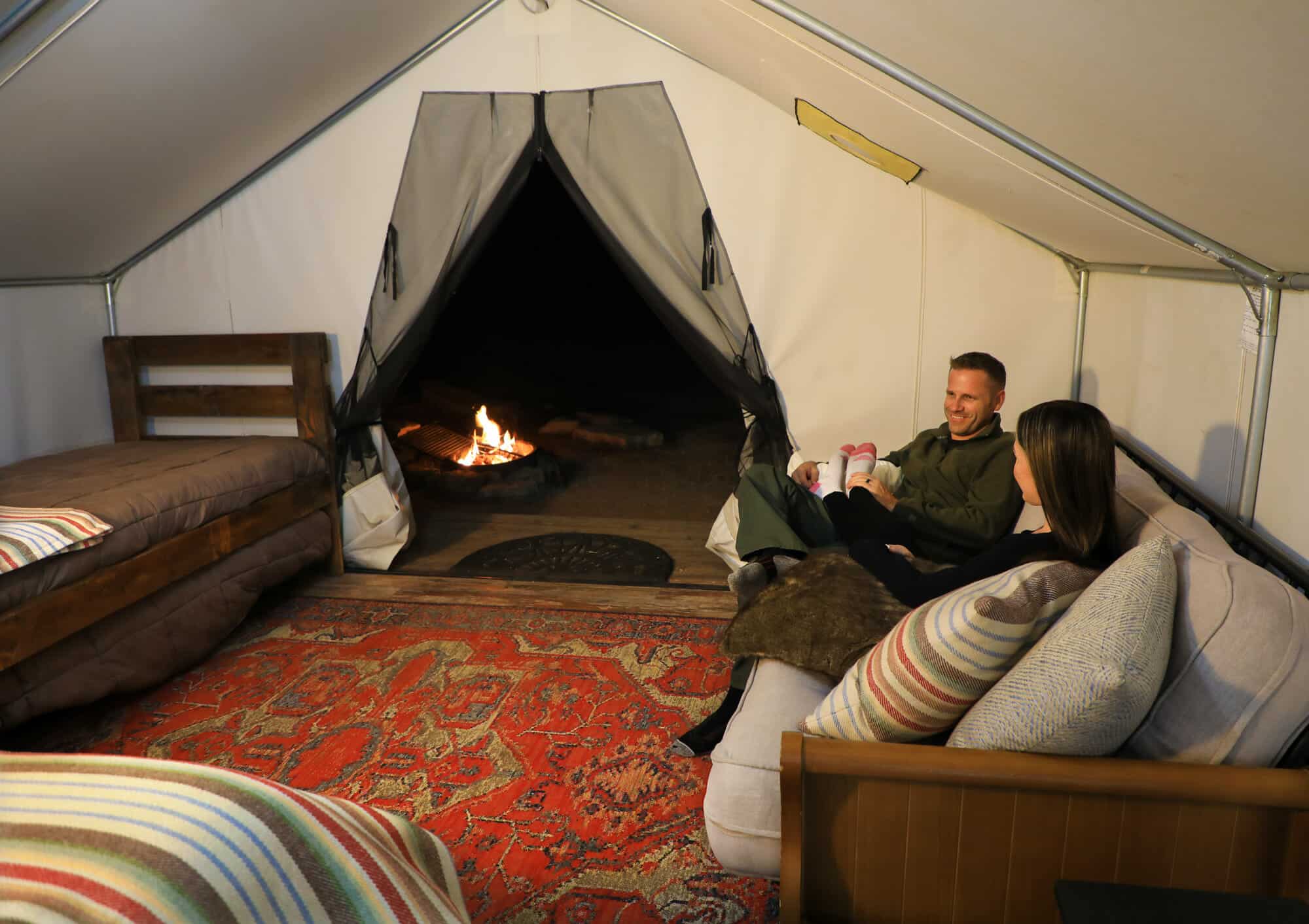 A couple relaxes inside a cozy tent with a fire pit visible outside.