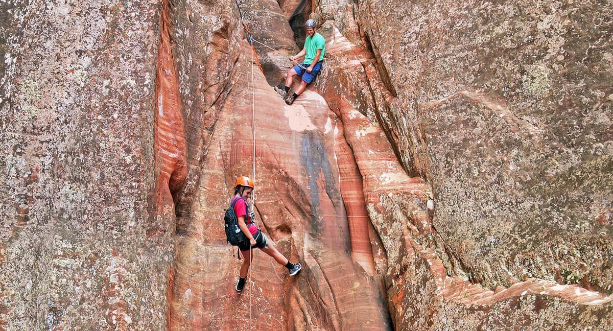 Two climbers descend a rocky canyon wall, one belaying while the other rappels down.