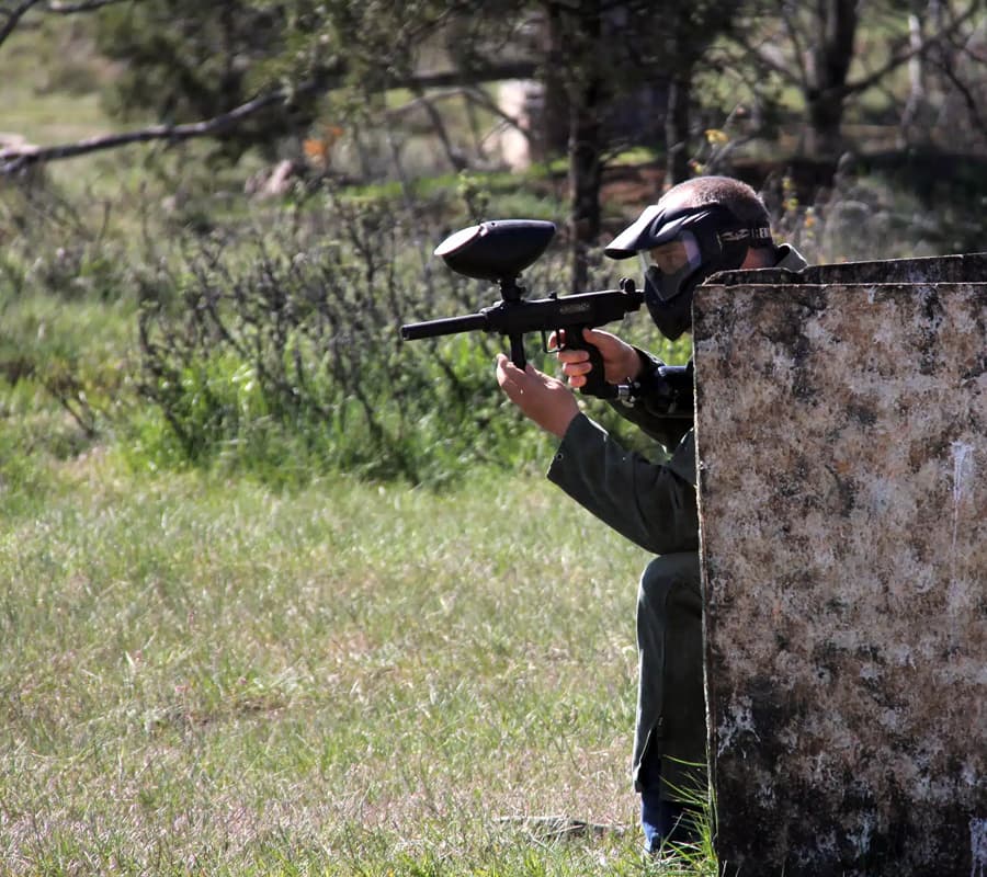 A person in a mask aims a paintball gun from behind a cover in a grassy field.
