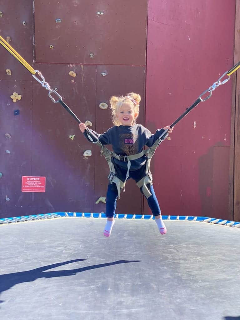 A smiling child enjoys bouncing on a trampoline while secured in harnesses.