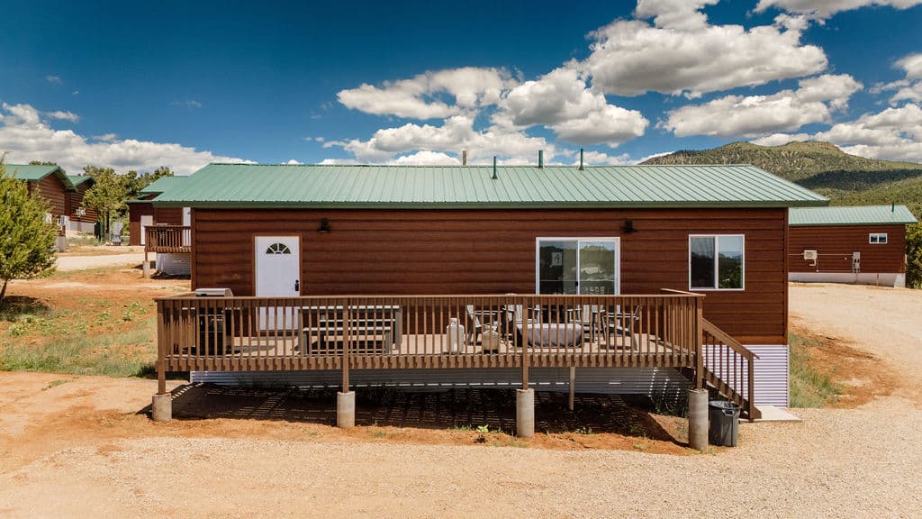 A brown wooden house with a green metal roof and a front deck, set in a grassy landscape under a blue sky with clouds.