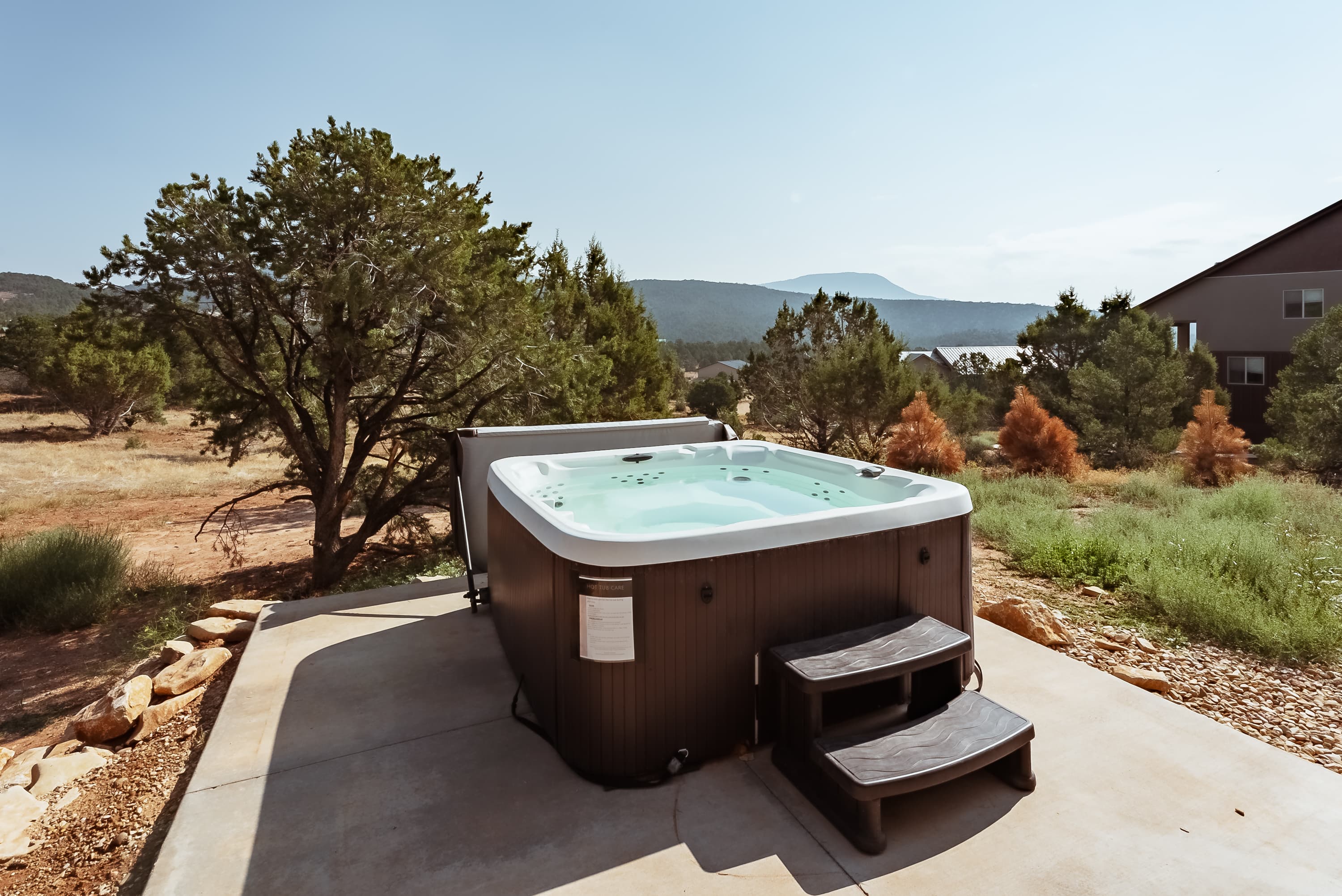 A hot tub sits on a patio surrounded by greenery and mountains under a clear sky.