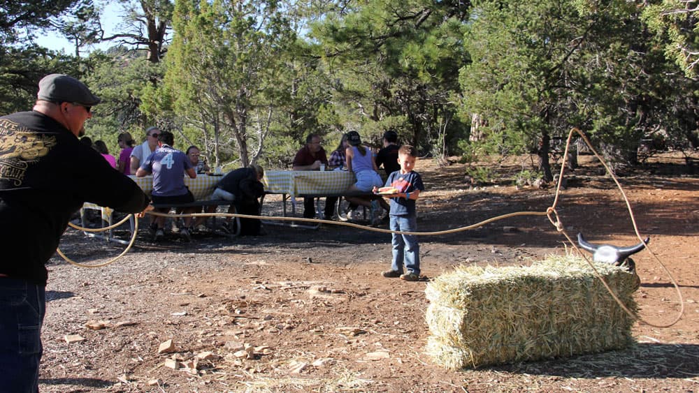 A man practices roping technique while a young boy watches near a hay bale, with a group seated at a table in the background.