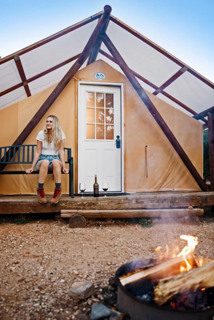A woman sits on a bench outside a glamping tent, with a campfire and wine glasses nearby.