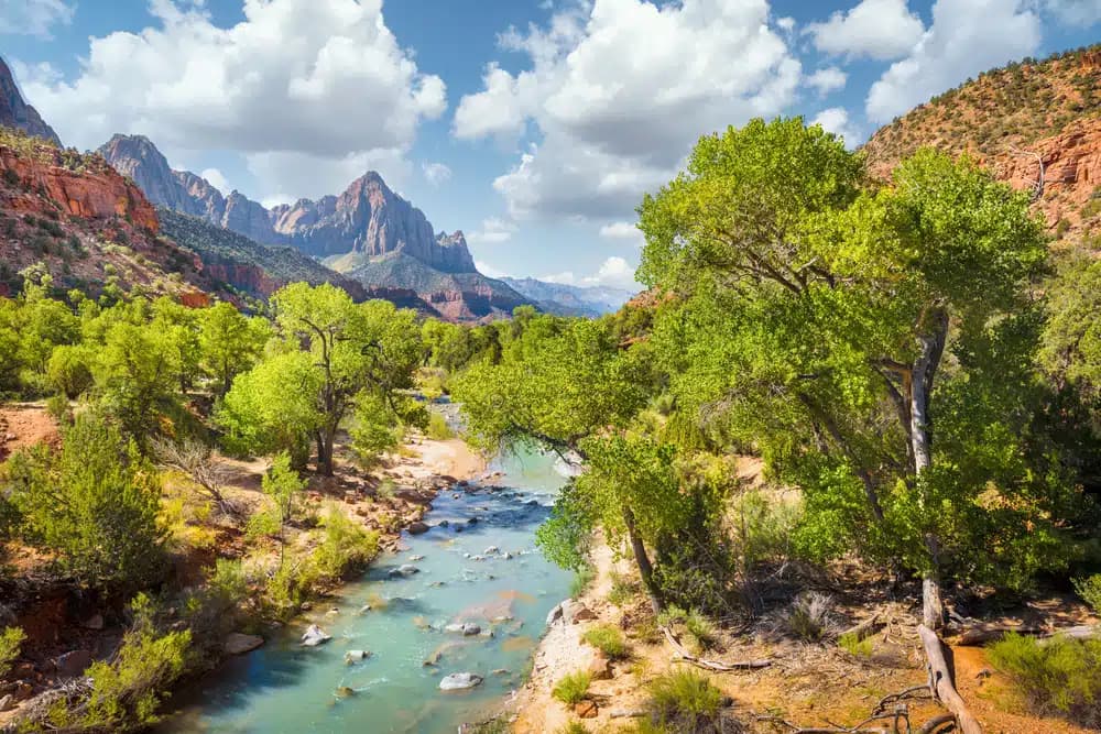 A serene river flows through lush greenery, framed by majestic mountains under a partly cloudy sky.