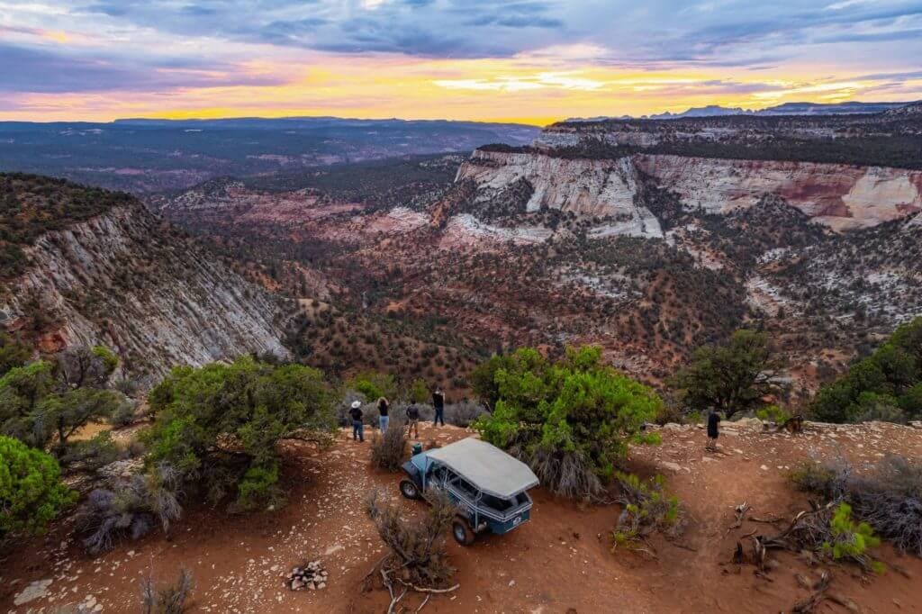 A group of people stand near a vintage blue vehicle, overlooking a vast canyon at sunset.
