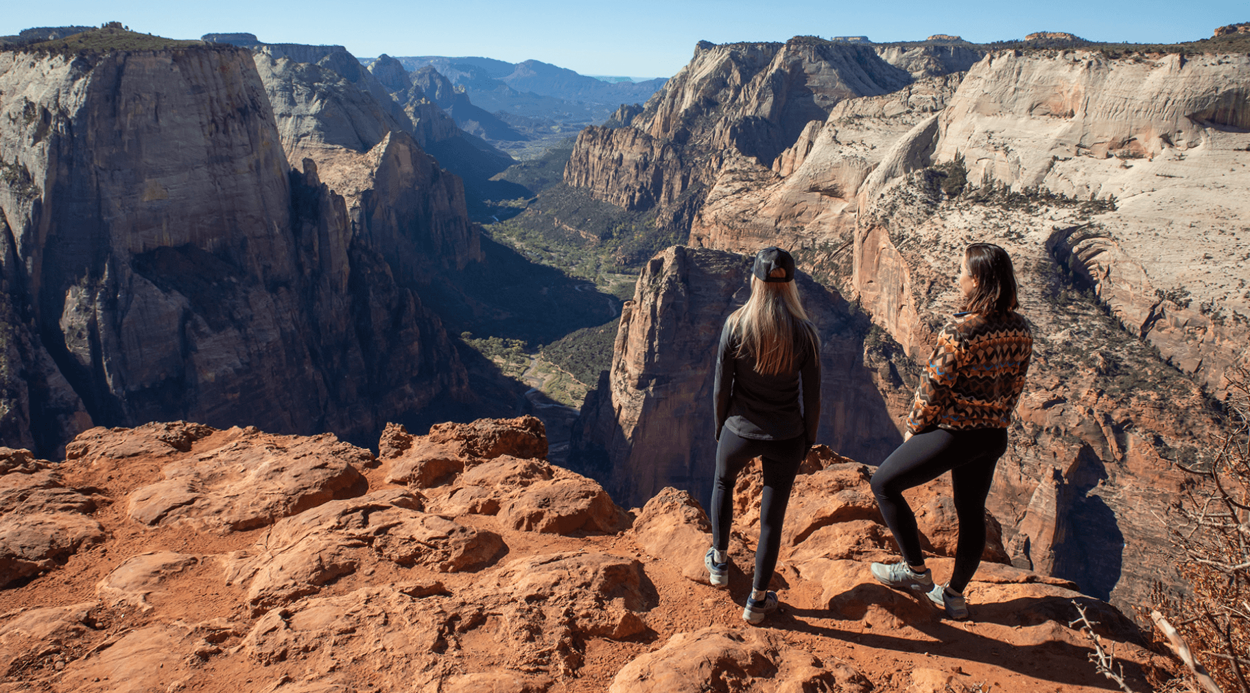 Two women stand on a rocky ledge overlooking a vast canyon landscape.