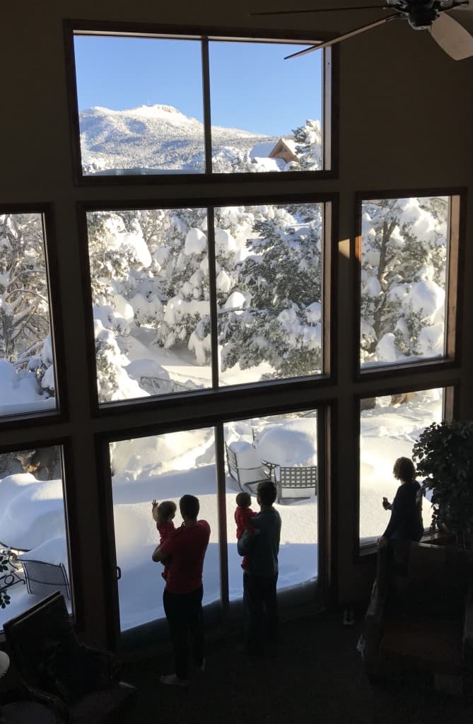 A family looks out of a large window at a snowy landscape and mountains.