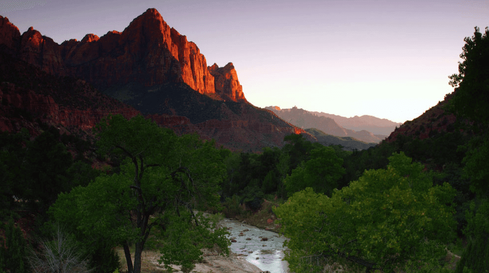 Sunlit red rock mountains overlook a winding river surrounded by lush greenery.