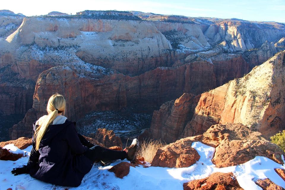 A person sits on a rocky ledge overlooking a snow-dusted canyon landscape.