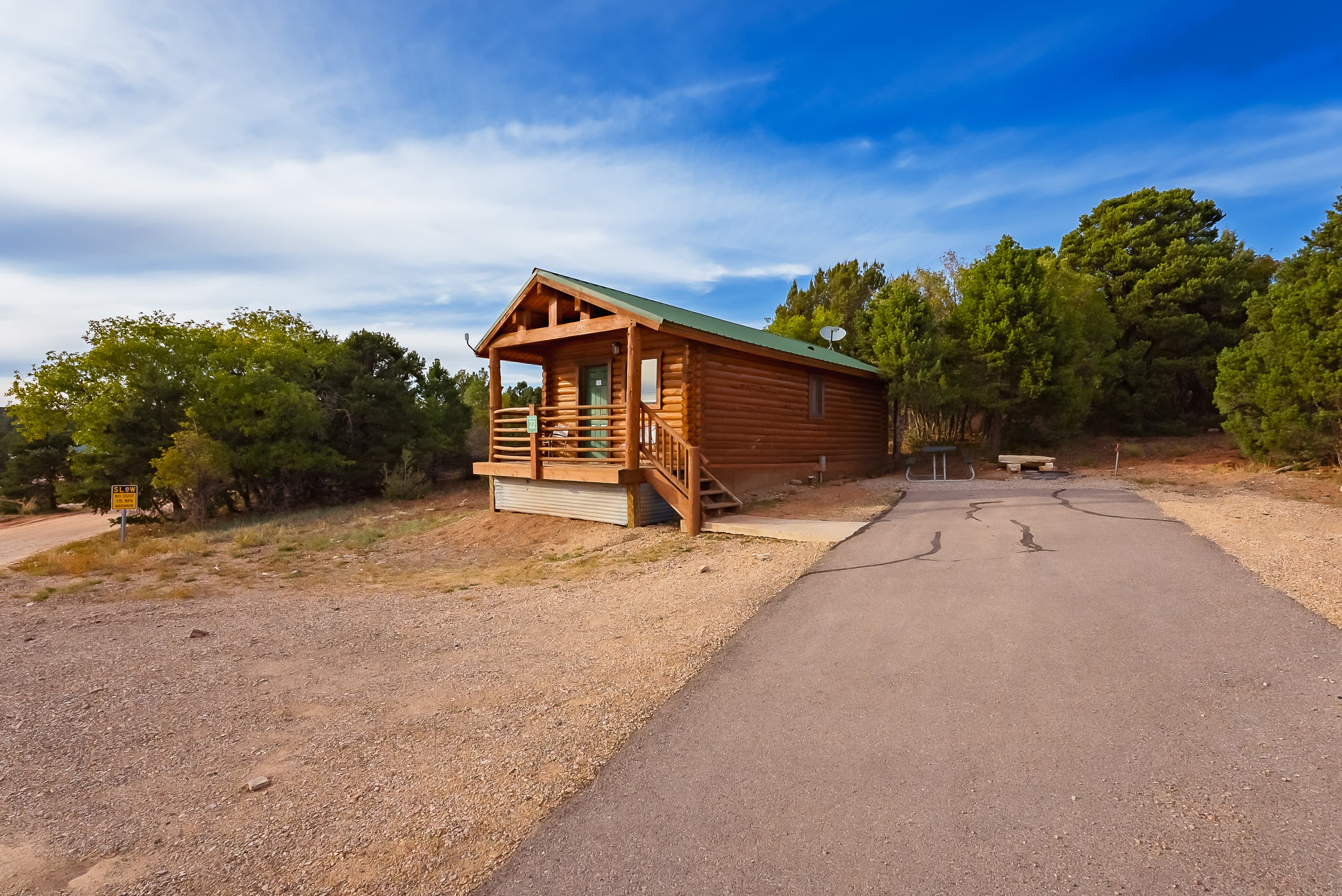 A rustic log cabin with a green roof set against a backdrop of trees and a clear blue sky.